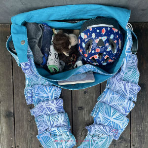Top view of the inside of the babywearing bag containing a large quantity of things: cloth nappies, a wool one-piece suit, a bike/trike helmet, a monkey plushy, a glasses case, a book, a reusable water bottle, a large wallet, a set of keys, a pack of tissues... and could still hold some more