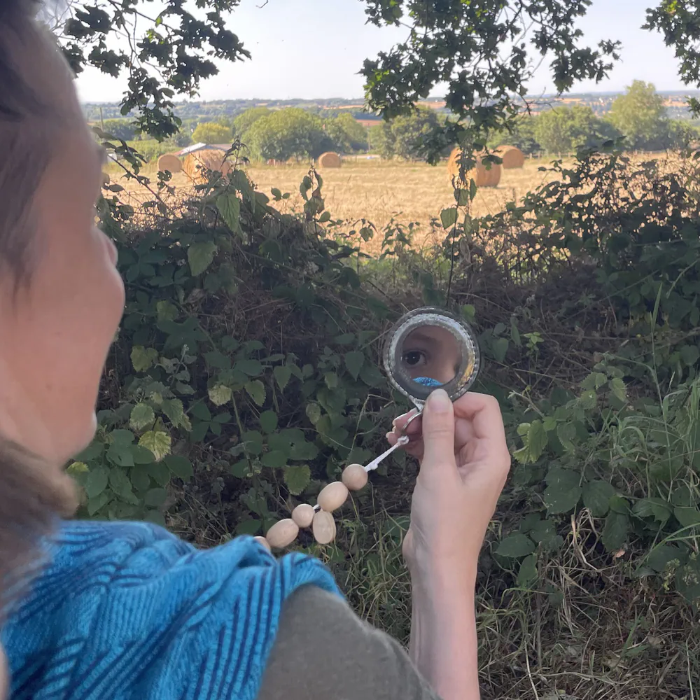 Une maman blanche regarde son bambin blanc dans un miroir portatif en cuir, l'enfant est porté dans une écharpe tissée main, ils sont à l'extérieur.